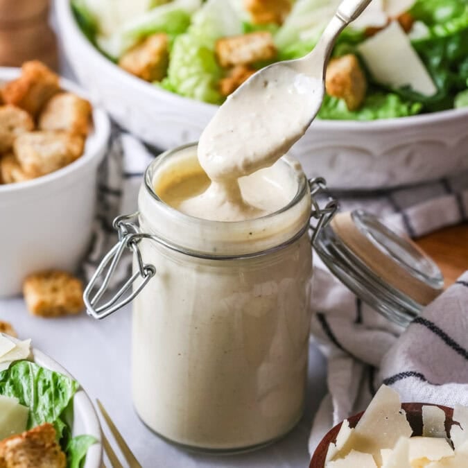 Spoon in a jar of homemade caesar dressing with salads in the background.