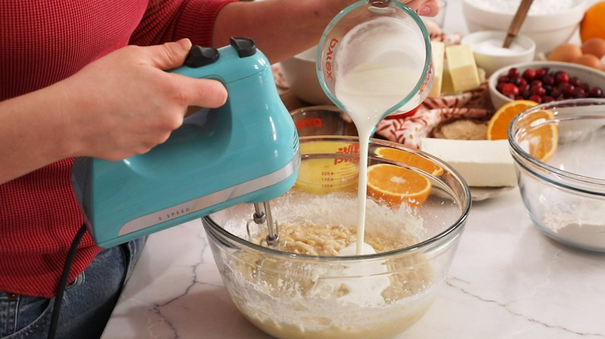 Buttermilk being poured into cranberry orange cake batter while mixing with a hand mixer.