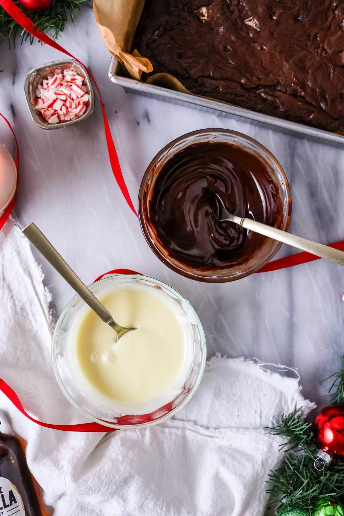 Two bowls of melted chocolate, one white chocolate and one dark, beside a pan of brownies.