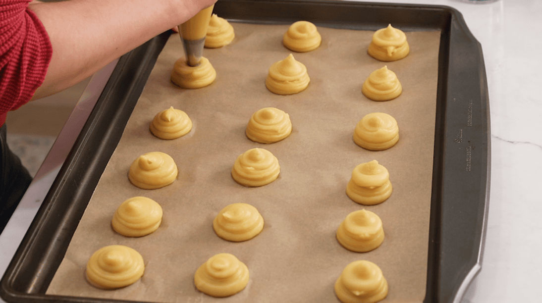Piping choux dough into mounds on a baking sheet.
