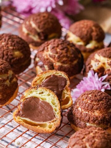 Chocolate choux au craquelin on a cooling rack with one choux bun cut in half to show its chocolate pastry cream center.