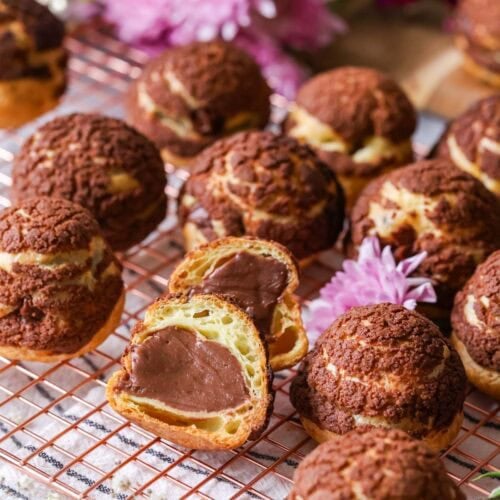 Chocolate choux au craquelin on a cooling rack with one choux bun cut in half to show its chocolate pastry cream center.