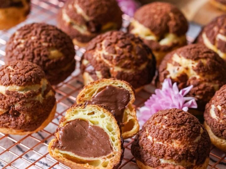 Chocolate choux au craquelin on a cooling rack with one choux bun cut in half to show its chocolate pastry cream center.