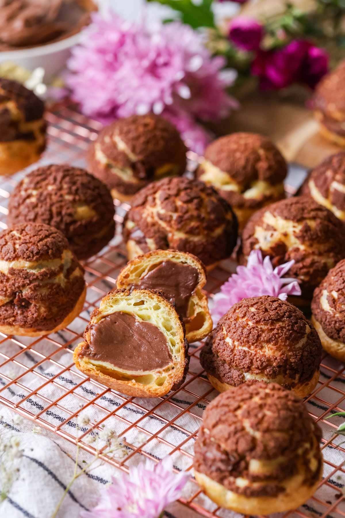 Chocolate choux au craquelin on a cooling rack with one choux bun cut in half to show its chocolate pastry cream center.