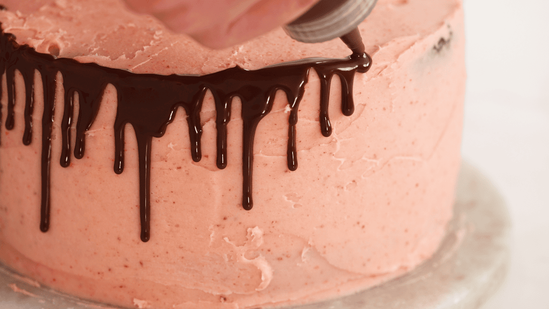 Chocolate ganache drip being drizzled around the edge of a pink frosted cake.