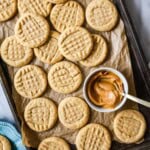 Overhead view of peanut butter cookies next to a bowl of peanut butter on a parchment lined baking sheet.