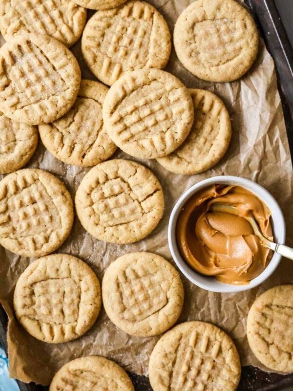 Overhead view of peanut butter cookies next to a bowl of peanut butter on a parchment lined baking sheet.
