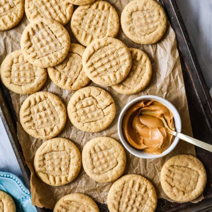 Overhead view of peanut butter cookies next to a bowl of peanut butter on a parchment lined baking sheet.