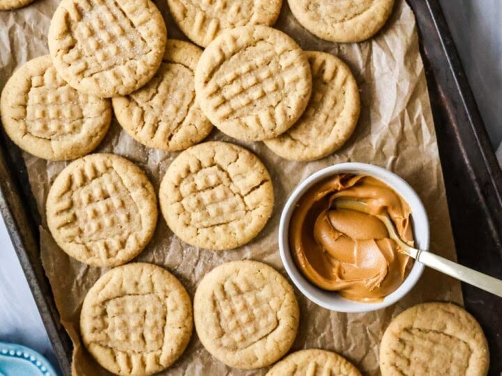 Overhead view of peanut butter cookies next to a bowl of peanut butter on a parchment lined baking sheet.