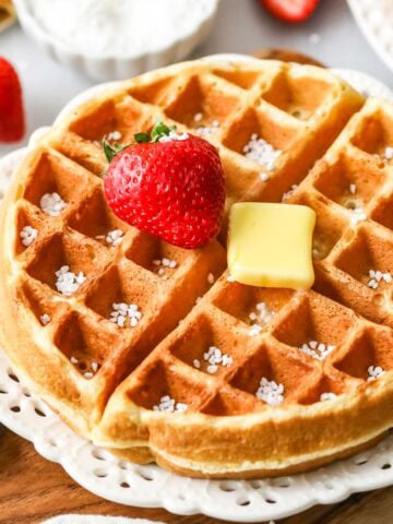 Sourdough waffle on a plate topped with a strawberry, a pat of butter, and Belgian pearl sugar.