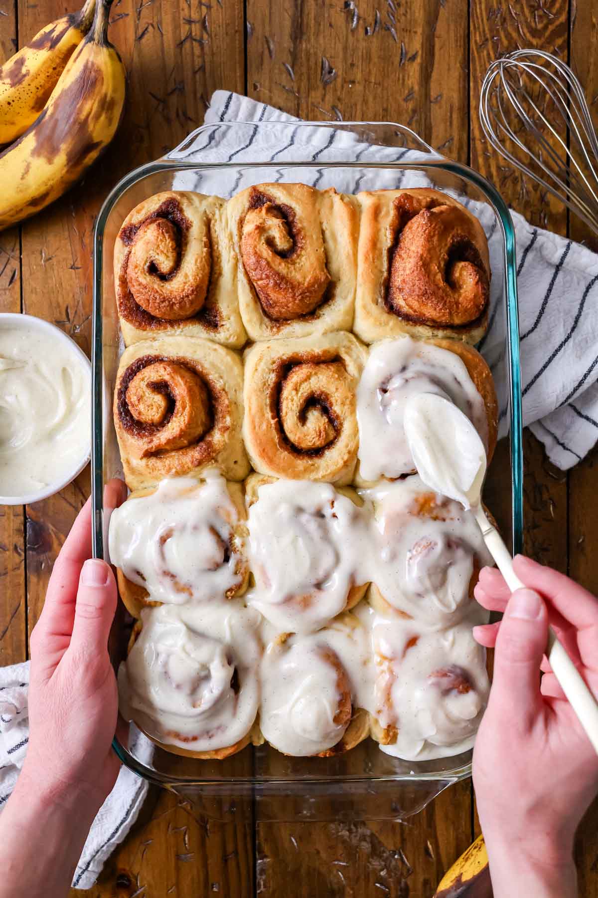 Overhead view of banana bread cinnamon rolls being frosted with cream cheese icing.