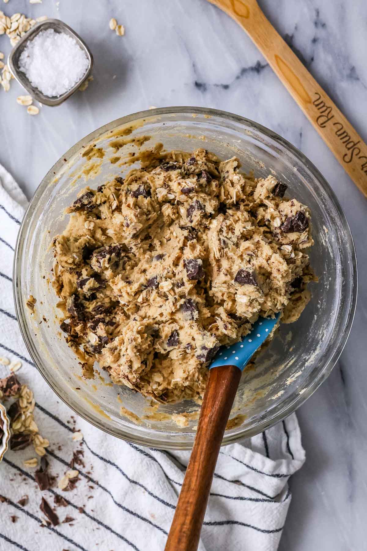 Glass mixing bowl with chocolate chip oatmeal cookie dough and a blue wood-handled spatula.