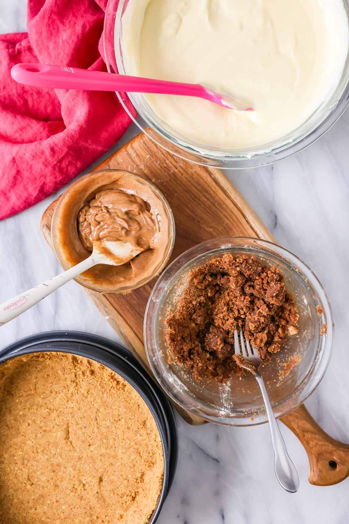 Bowls of components for a cinnamon roll cheesecake including cheesecake batter, graham cracker crust, cinnamon sugar paste, and cinnamon sugar crumbles.
