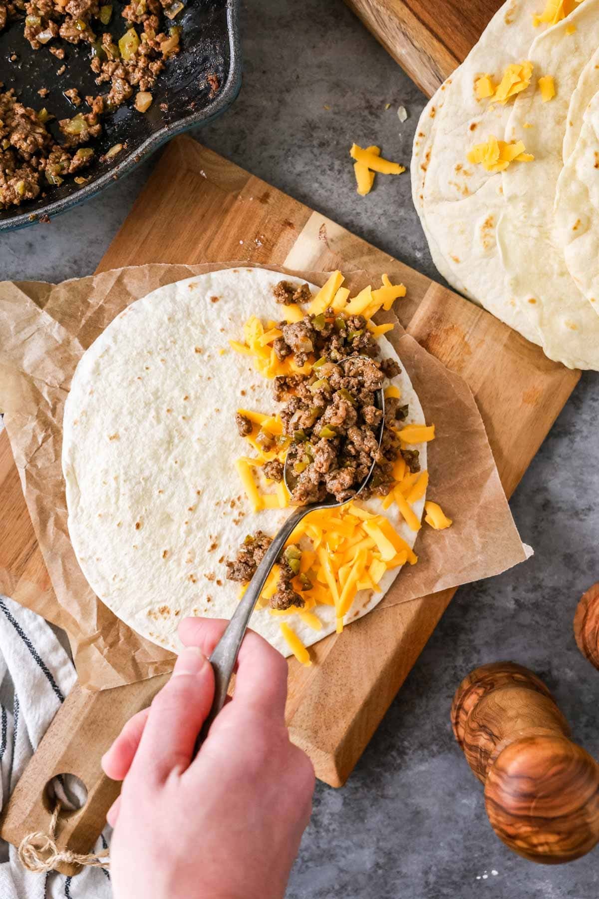 Seasoned ground beef being spooned over shredded cheese on a flour tortilla.