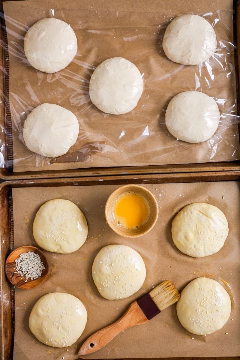 Overhead view of two trays of unbaked burger buns, one covered in plastic wrap and the other brushed with egg wash and sesame seeds.
