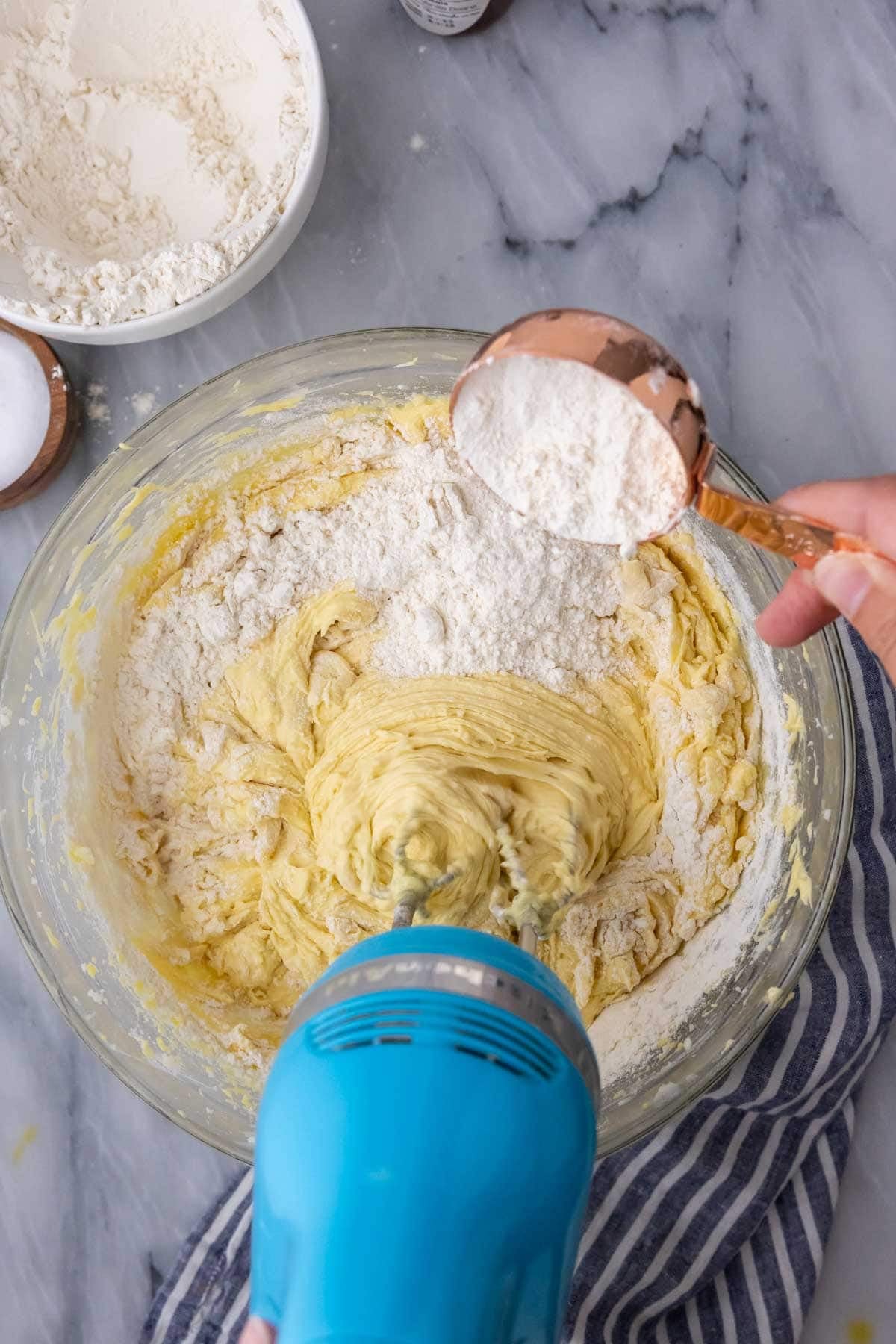 Stirring flour into pound cake batter in a large glass mixing bowl with a blue mixer.
