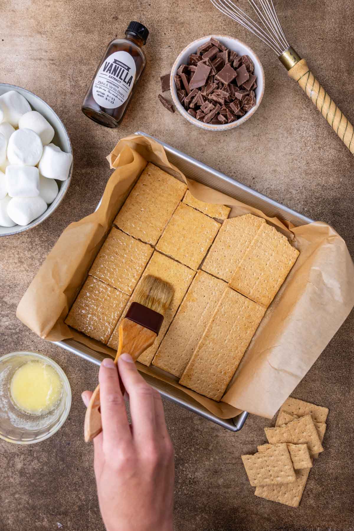 Overhead view of butter being brushed over graham crackers in a square baking pan.