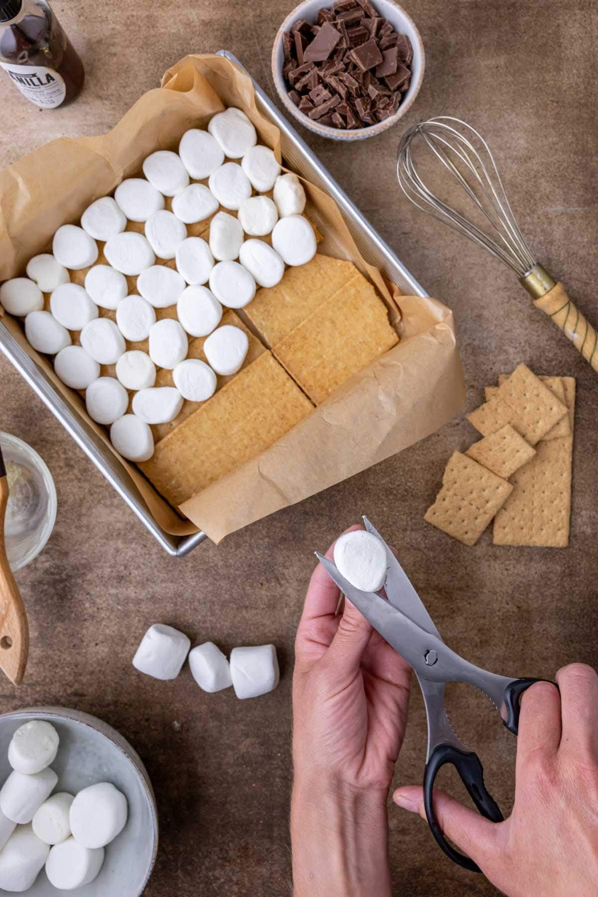 Overhead view of marshmallows being cut in half and layered over graham crackers in a square baking pan.
