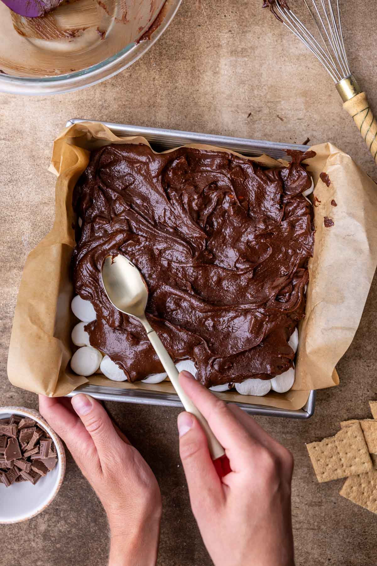 Overhead view of a spoon being used to spread brownie batter over a layer of marshmallows.