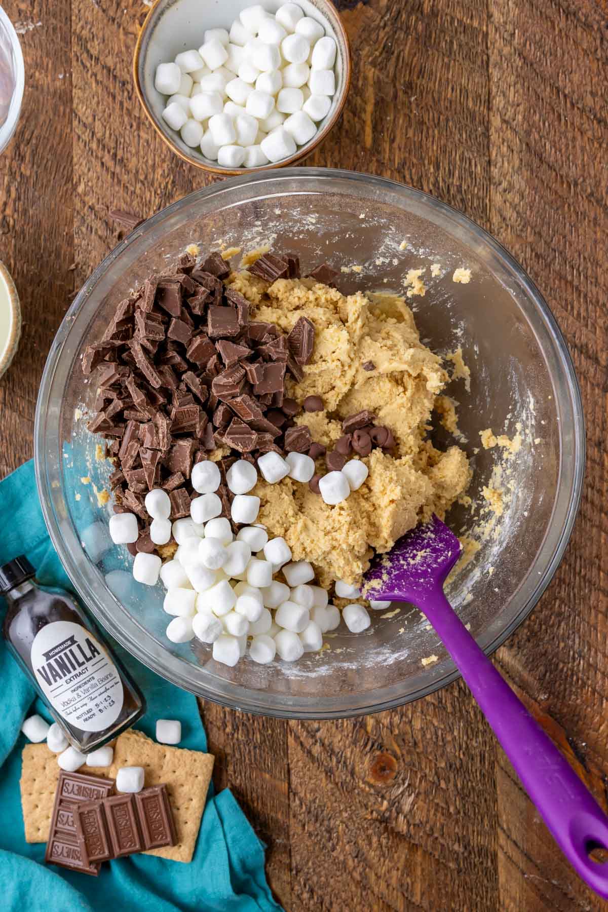 Overhead of clear bowl with cookie dough, milk chocolate chips, and marshmallows being mixed together to make smores cookies