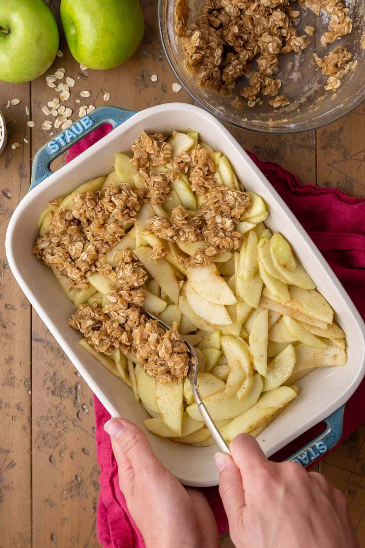 Sprinkling oat crisp mixture over apples layered into a blue baking dish.