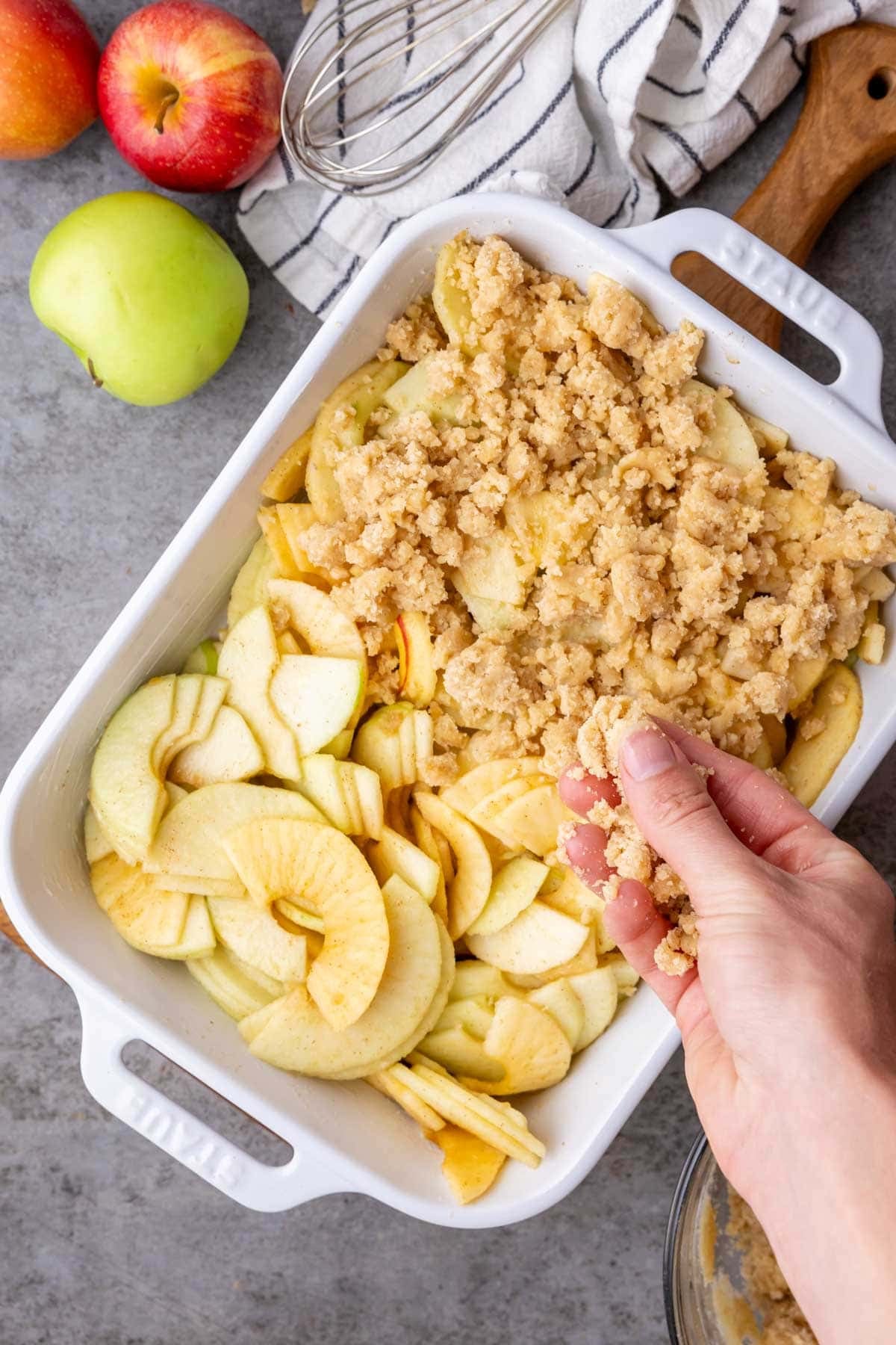 Hands sprinkling a crumble topping over spiced apples in a baking dish.