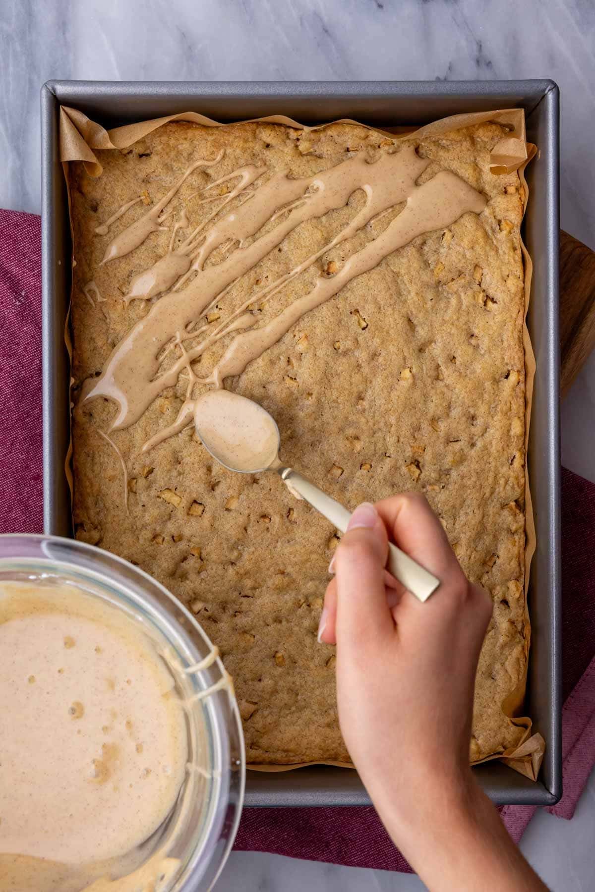 Drizzling a spiced glaze over a pan of blondies made with apples.