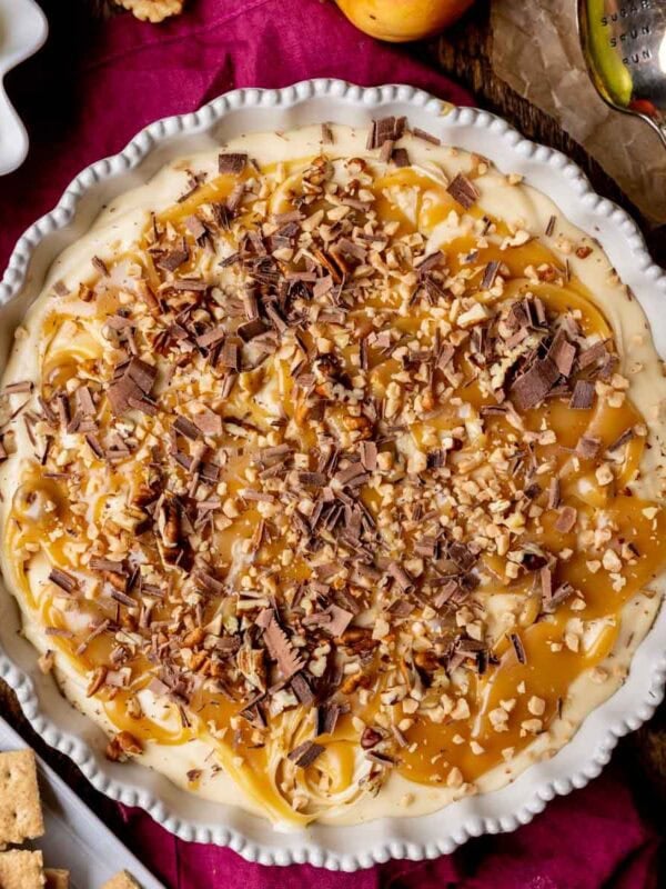 Overhead view of caramel apple cheesecake dip topped with caramel, pecans, shaved chocolate, and toffee in a round serving dish.