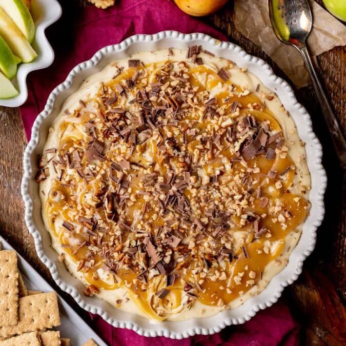 Overhead view of caramel apple cheesecake dip topped with caramel, pecans, shaved chocolate, and toffee in a round serving dish.