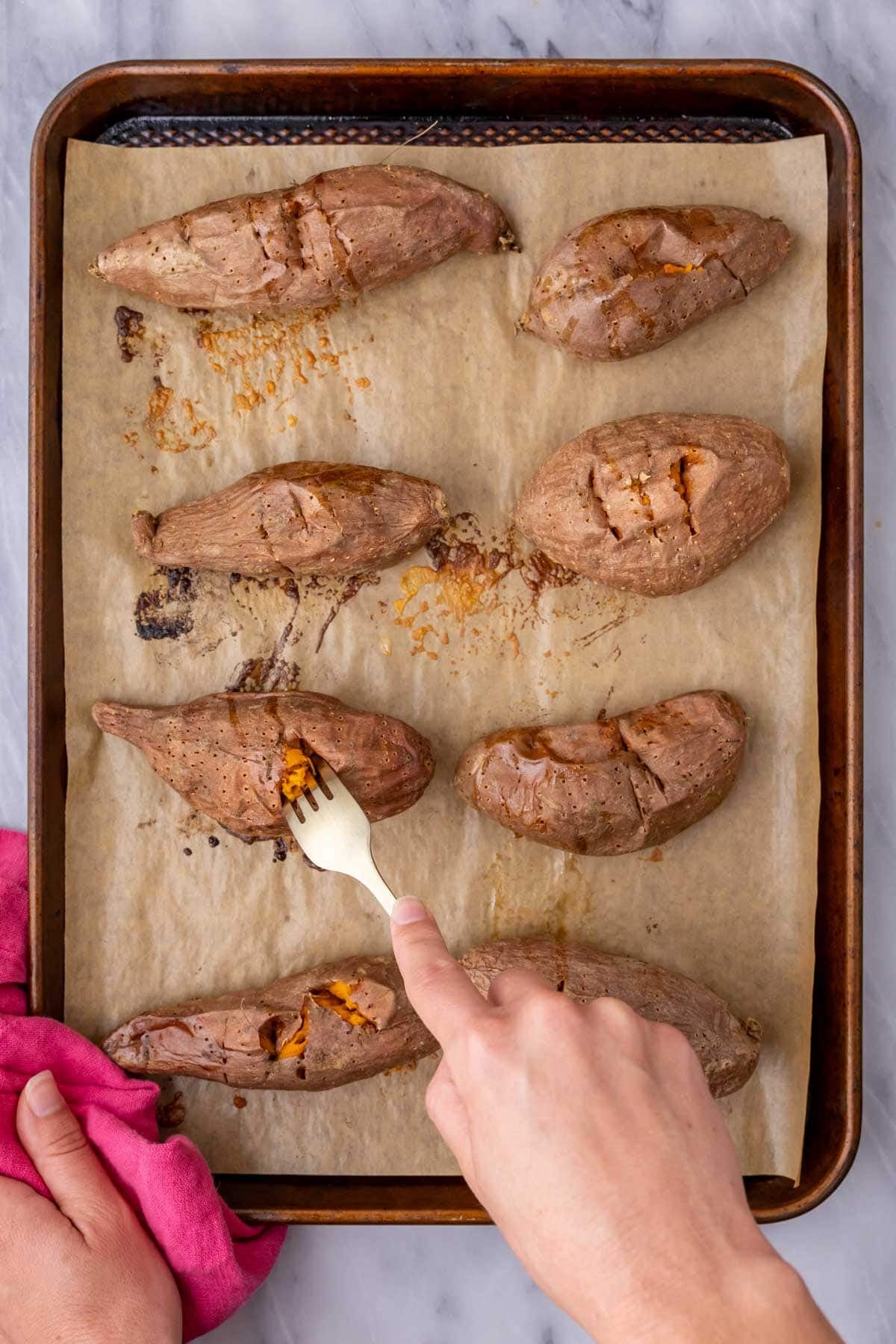 Roasted sweet potatoes on a baking sheet being pierced with a gold fork.