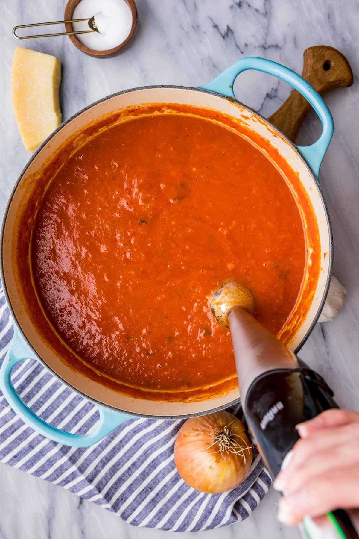 Overhead of tomato soup in a blue pot being pureed with a stick immersion blender.