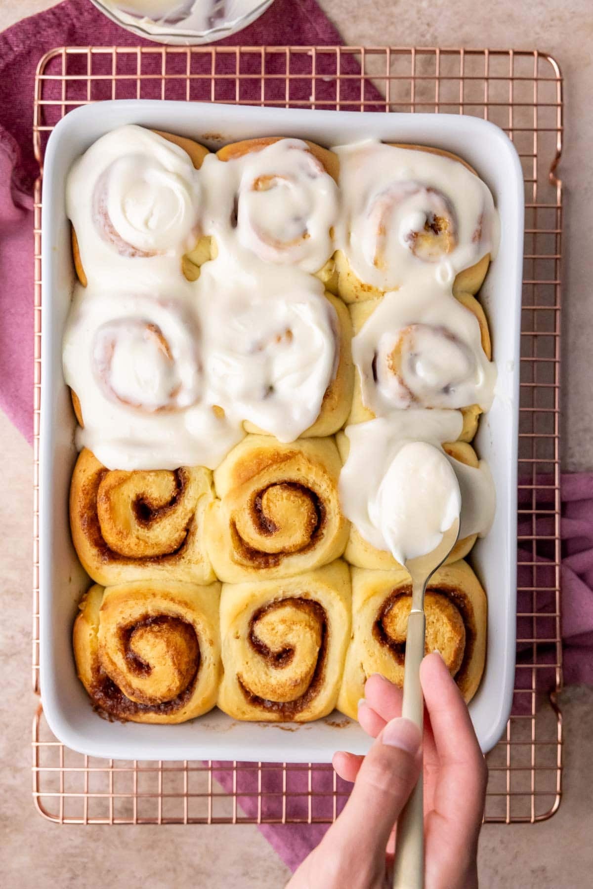 Cream cheese frosting being spread over warm cinnamon rolls in a white dish.