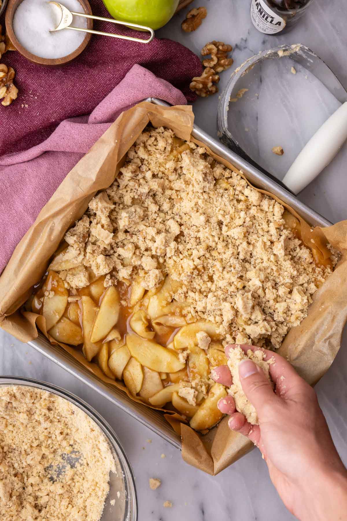 Hands scattering a crumble topping over an apple filling.