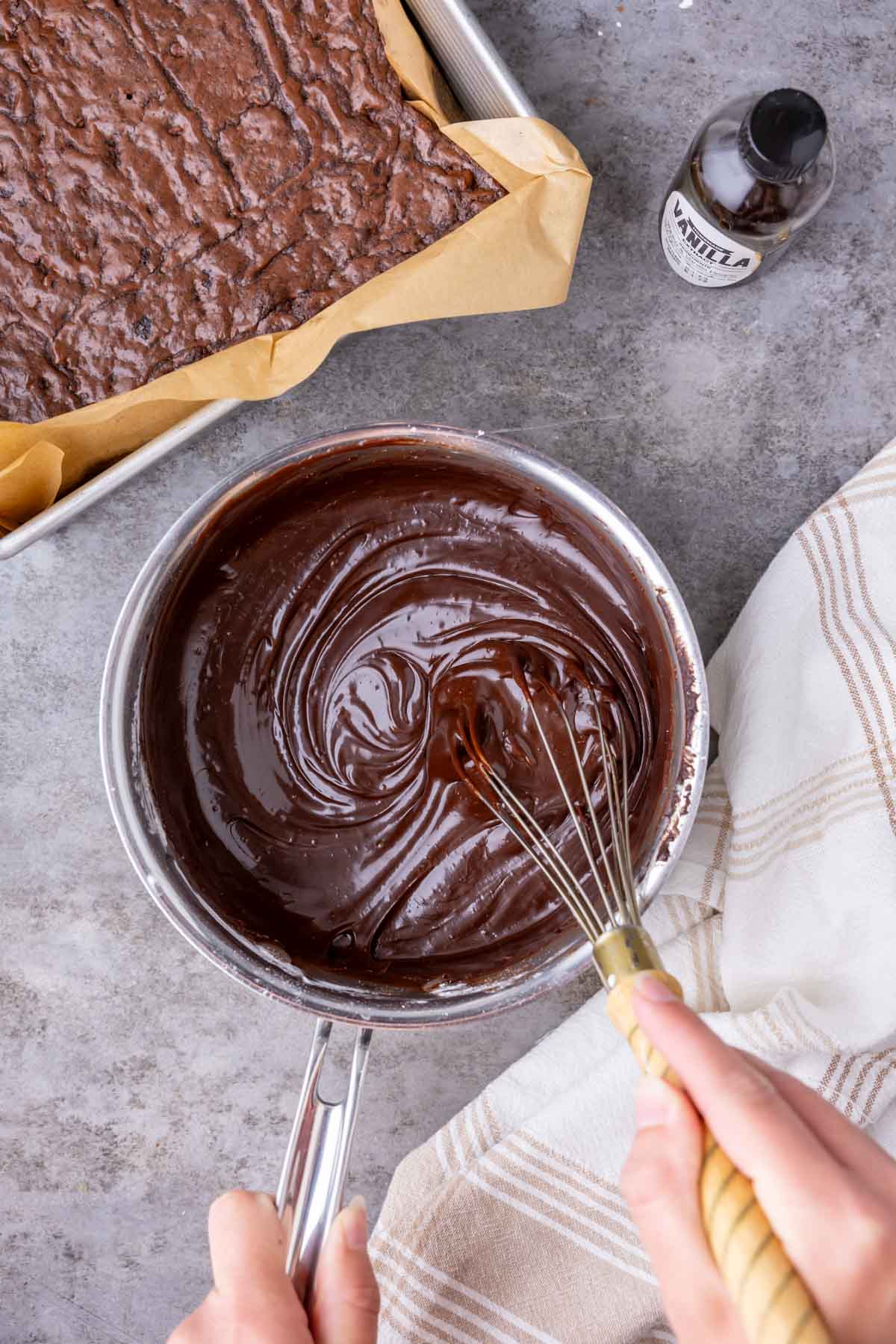 Overhead view of a fudge frosting being whisked together in a saucepan beside a pan of brownies.